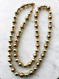 Vintage gold and bronze glass beaded flapper necklace laid on a white marble surface, featuring alternating reflective gold beads and matte bronze pearls with gold-tone spacers.