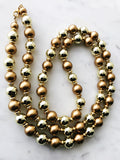 Vintage gold and bronze glass beaded flapper necklace laid on a white marble surface, featuring alternating reflective gold beads and matte bronze pearls with gold-tone spacers.