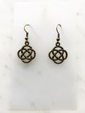 A pair of antique bronze Celtic knot dangle earrings displayed on a white card over a light marble surface.