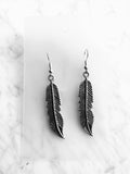 A pair of silver-toned feather drop earrings with detailed textured barbs, displayed on a clear acrylic card over a white marble background.