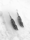 A pair of silver-toned feather drop earrings with detailed textured barbs, displayed on a clear acrylic card over a white marble background.