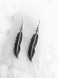 A pair of silver-toned feather drop earrings with detailed textured barbs, displayed on a clear acrylic card over a white marble background.