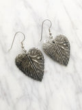  A pair of silver-coloured leaf-shaped earrings. Each earring has a detailed leaf design with visible veins and a textured surface. The earrings hang from simple silver hooks, showcasing a nature-inspired jewellery design.