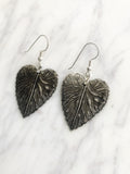  A pair of silver-coloured leaf-shaped earrings. Each earring has a detailed leaf design with visible veins and a textured surface. The earrings hang from simple silver hooks, showcasing a nature-inspired jewellery design.
