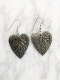 A pair of silver-coloured leaf-shaped earrings. Each earring has a detailed leaf design with visible veins and a textured surface. The earrings hang from simple silver hooks, showcasing a nature-inspired jewellery design.