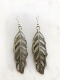 A pair of silver-toned drop earrings shaped like stylized feathers, displayed on a light marble surface.