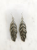 A pair of silver-toned feather drop earrings with detailed engraved patterns, displayed on a white marble surface.
