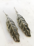 A pair of silver-toned feather drop earrings with detailed wire-like feather patterns, displayed on a light marble surface.