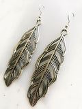 A pair of silver-toned feather drop earrings with detailed cutout leaf-like patterns and fishhook ear wires, displayed on a light marble surface.