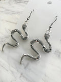 A pair of silver snake drop earrings with textured scales and fishhook wires, displayed on a white card over a white marble surface.