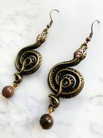 Pair of ornate winding serpent shape earrings, antique bronze tone with vintage crystals and Indian Agate beads on a marble background