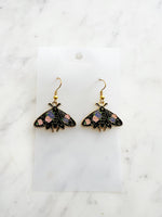Kawaii Black moth-shaped earrings with floral details on a white card and a marble background