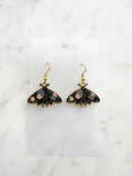 Kawaii Black moth-shaped earrings with floral details on a white card and a marble background