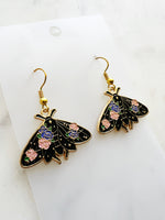 Kawaii Black moth-shaped earrings with floral details on a white card and a marble background