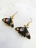Kawaii Black moth-shaped earrings with floral details on a white card and a marble background