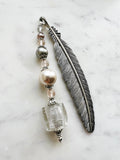 Side view of a Handmade silver feather bookmark with neutral-toned repurposed vintage beads, including a clear foil glass cube, pearls, and crystals, displayed on a marble background.