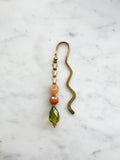 Handmade brass bookmark with warm-toned repurposed vintage beads in peach, orange, and olive green on a wavy brass hook, displayed on a marble background.