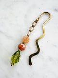 side view of a Handmade brass bookmark with warm-toned repurposed vintage beads in peach, orange, and olive green on a wavy brass hook, displayed on a marble background.