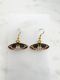 Pair of Kawaii enamel moth dangle earrings featuring rust-red wings with black and white detailing, outlined in gold-tone metal with gold ear hooks, displayed on a marble background.