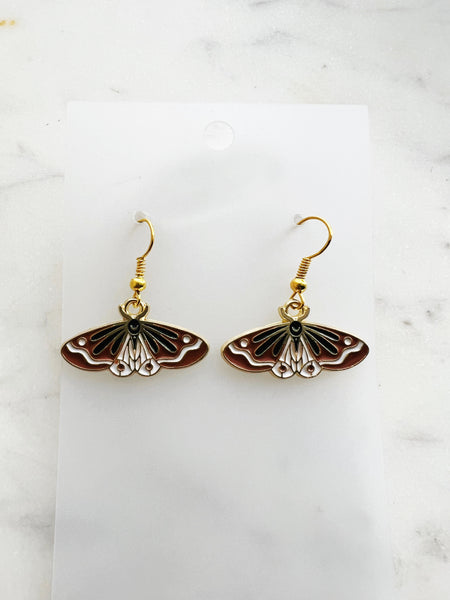Pair of Kawaii enamel moth dangle earrings featuring rust-red wings with black and white detailing, outlined in gold-tone metal with gold ear hooks, displayed on a marble background.