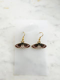 Pair of Kawaii enamel moth dangle earrings featuring rust-red wings with black and white detailing, outlined in gold-tone metal with gold ear hooks, displayed on a marble background.