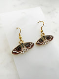 Side view of a Pair of Kawaii enamel moth dangle earrings featuring rust-red wings with black and white detailing, outlined in gold-tone metal with gold ear hooks, displayed on a marble background.