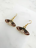 Side view of a Pair of Kawaii enamel moth dangle earrings featuring rust-red wings with black and white detailing, outlined in gold-tone metal with gold ear hooks, displayed on a marble background.