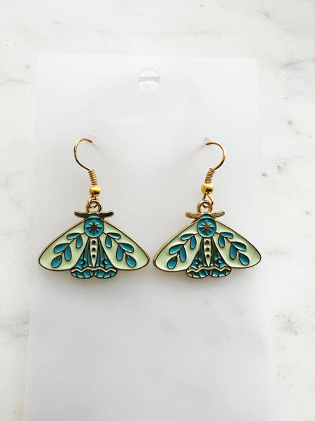 Pair of Kawaii enamel moth dangle earrings featuring pastel mint wings with teal botanical detailing, outlined in gold-tone metal with gold ear hooks, displayed on a marble background.