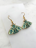 Side view of a Pair of Kawaii enamel moth dangle earrings featuring pastel mint wings with teal botanical detailing, outlined in gold-tone metal with gold ear hooks, displayed on a marble background.