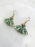 Side view of a Pair of Kawaii enamel moth dangle earrings featuring pastel mint wings with teal botanical detailing, outlined in gold-tone metal with gold ear hooks, displayed on a marble background.