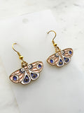 Side view of a pair of Kawaii enamel moth dangle earrings featuring pastel blush wings with periwinkle and white accents, decorated with crescent moons, stars and dotted patterns, outlined in gold-tone metal with gold ear hooks, displayed on a marble background.