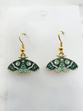Pair of Kawaii enamel moth dangle earrings featuring teal and mint wings with crescent moon, star and decorative patterns, outlined in gold-tone metal with gold ear hooks, displayed on a marble background.