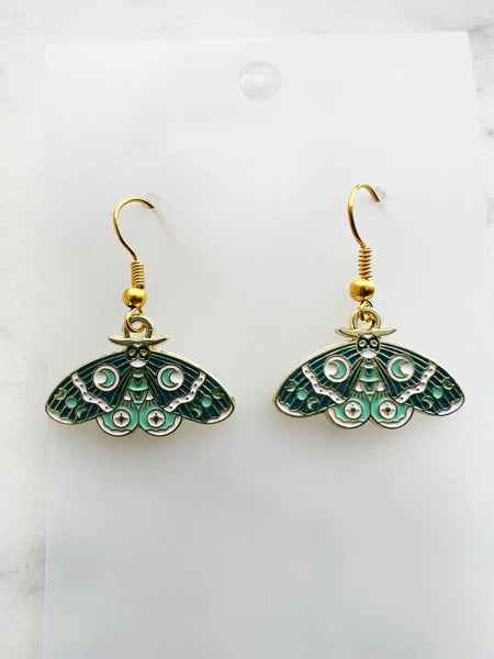 Pair of Kawaii enamel moth dangle earrings featuring teal and mint wings with crescent moon, star and decorative patterns, outlined in gold-tone metal with gold ear hooks, displayed on a marble background.