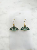 Pair of Kawaii enamel moth dangle earrings featuring teal and mint wings with crescent moon, star and decorative patterns, outlined in gold-tone metal with gold ear hooks, displayed on a marble background.