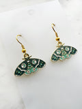 Side view of a pair of Kawaii enamel moth dangle earrings featuring teal and mint wings with crescent moon, star and decorative patterns, outlined in gold-tone metal with gold ear hooks, displayed on a marble background.