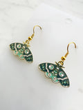 side view of a pair of Kawaii enamel moth dangle earrings featuring teal and mint wings with crescent moon, star and decorative patterns, outlined in gold-tone metal with gold ear hooks, displayed on a marble background.
