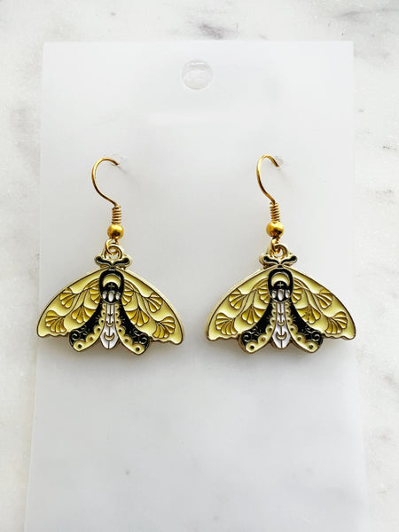 Pair of Kawaii enamel moth dangle earrings featuring pastel yellow wings with black detailing and decorative leaf motifs, outlined in gold-tone metal with gold ear hooks, displayed on a marble background.