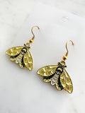 side view pf a pair of Kawaii enamel moth dangle earrings featuring pastel yellow wings with black detailing and decorative leaf motifs, outlined in gold-tone metal with gold ear hooks, displayed on a marble background.