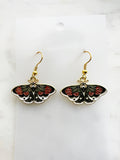 Pair of Kawaii enamel moth dangle earrings featuring black wings with red flowers, green leaves, and white accents, outlined in gold-tone metal with gold ear hooks, displayed against a marble background.