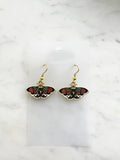 Pair of Kawaii enamel moth dangle earrings featuring black wings with red flowers, green leaves, and white accents, outlined in gold-tone metal with gold ear hooks, displayed against a marble background.