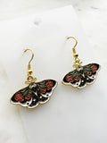 Side view pair of Kawaii enamel moth dangle earrings featuring black wings with red flowers, green leaves, and white accents, outlined in gold-tone metal with gold ear hooks, displayed against a marble background.
