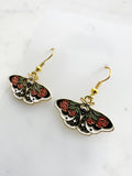 Side view pair of Kawaii enamel moth dangle earrings featuring black wings with red flowers, green leaves, and white accents, outlined in gold-tone metal with gold ear hooks, displayed against a marble background.
