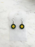 Pair of Kawaii enamel flower dangle earrings featuring green petals with a yellow centre, outlined in silver-tone metal with silver ear hooks, displayed on a marble background.