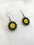 Side view of a Pair of Kawaii enamel flower dangle earrings featuring green petals with a yellow centre, outlined in silver-tone metal with silver ear hooks, displayed on a marble background.