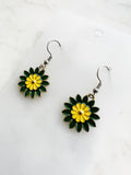 Side view of a Pair of Kawaii enamel flower dangle earrings featuring green petals with a yellow centre, outlined in silver-tone metal with silver ear hooks, displayed on a marble background.
