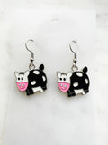 Pair of Kawaii enamel cow dangle earrings featuring black-and-white cows with pink snouts, outlined in silver-tone metal with silver ear hooks, displayed on a marble background.