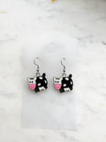 Pair of Kawaii enamel cow dangle earrings featuring black-and-white cows with pink snouts, outlined in silver-tone metal with silver ear hooks, displayed on a marble background.