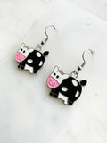 Side view of a Pair of Kawaii enamel cow dangle earrings featuring black-and-white cows with pink snouts, outlined in silver-tone metal with silver ear hooks, displayed on a marble background.