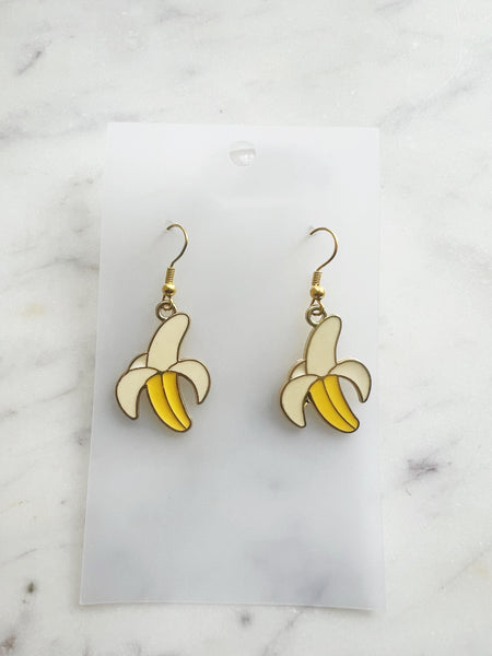 Pair of Kawaii enamel banana dangle earrings featuring yellow bananas with cream peel detailing, outlined in gold-tone metal with gold ear hooks, displayed on a marble background.
