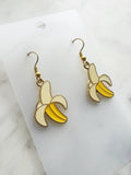 Side view of a Pair of Kawaii enamel banana dangle earrings featuring yellow bananas with cream peel detailing, outlined in gold-tone metal with gold ear hooks, displayed on a marble background